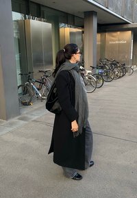 Juodas ilgas paltas, pilka šalikėlis ir tamsios kelnės. Lygus juodas krepšys, akiniai nuo saulės. Fone matomi dviračiai ir modernių pastatų įėjimas.