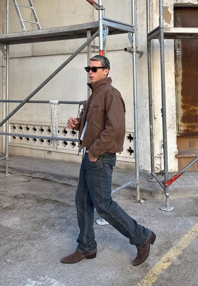 Hombre con chaqueta marrón, gafas de sol, vaqueros y botas marrones caminando junto a andamios metálicos en una calle de hormigón.