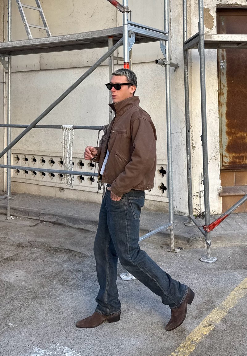 Man som bär brun jacka, solglasögon, jeans och bruna stövlar som går förbi metallställning på en betonggata.