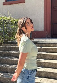 Miętowy obcisły t-shirt z krótkim rękawem zestawiony z szortami dżinsowymi o wysokim stanie, stojący na kamiennych schodach z rozmytym tłem.