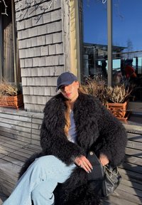 Femme portant un manteau noir en fausse fourrure et une casquette bleu marine, assise sur un banc en bois, tenant un sac à main noir, avec des pots de plantes et une fenêtre derrière elle.