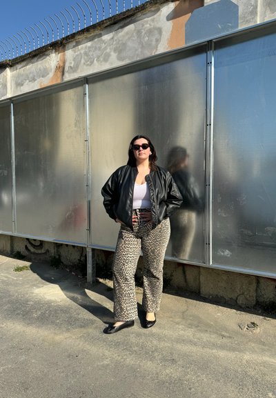 Chaqueta bomber de cuero negro sobre una blusa blanca, combinada con pantalones acampanados de leopardo beige y zapatos planos negros, de pie contra un fondo metálico.