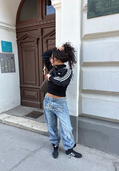 Joven mujer con chaqueta negra de Adidas y vaqueros anchos bordados está de pie en la acera ajustando su coleta rizada junto a una puerta marrón y la pared blanca de un edificio.