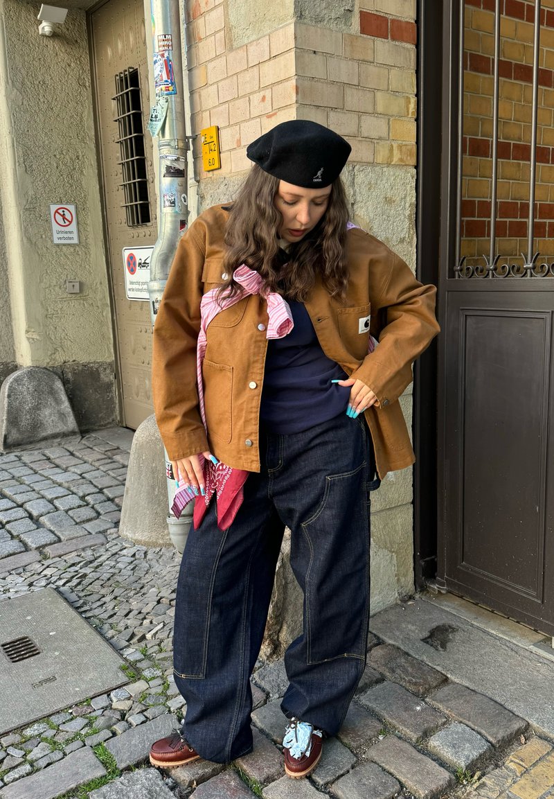 Jeune femme en veste marron, béret noir, jean ample et sombre, debout dans une rue pavée près d'un bâtiment d'angle avec des murs en brique et en pierre.