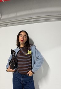 Femme aux longs cheveux bruns portant un cardigan bleu, un chemisier rayé marron et un jean, tenant un sac à main en cuir noir contre un mur blanc.