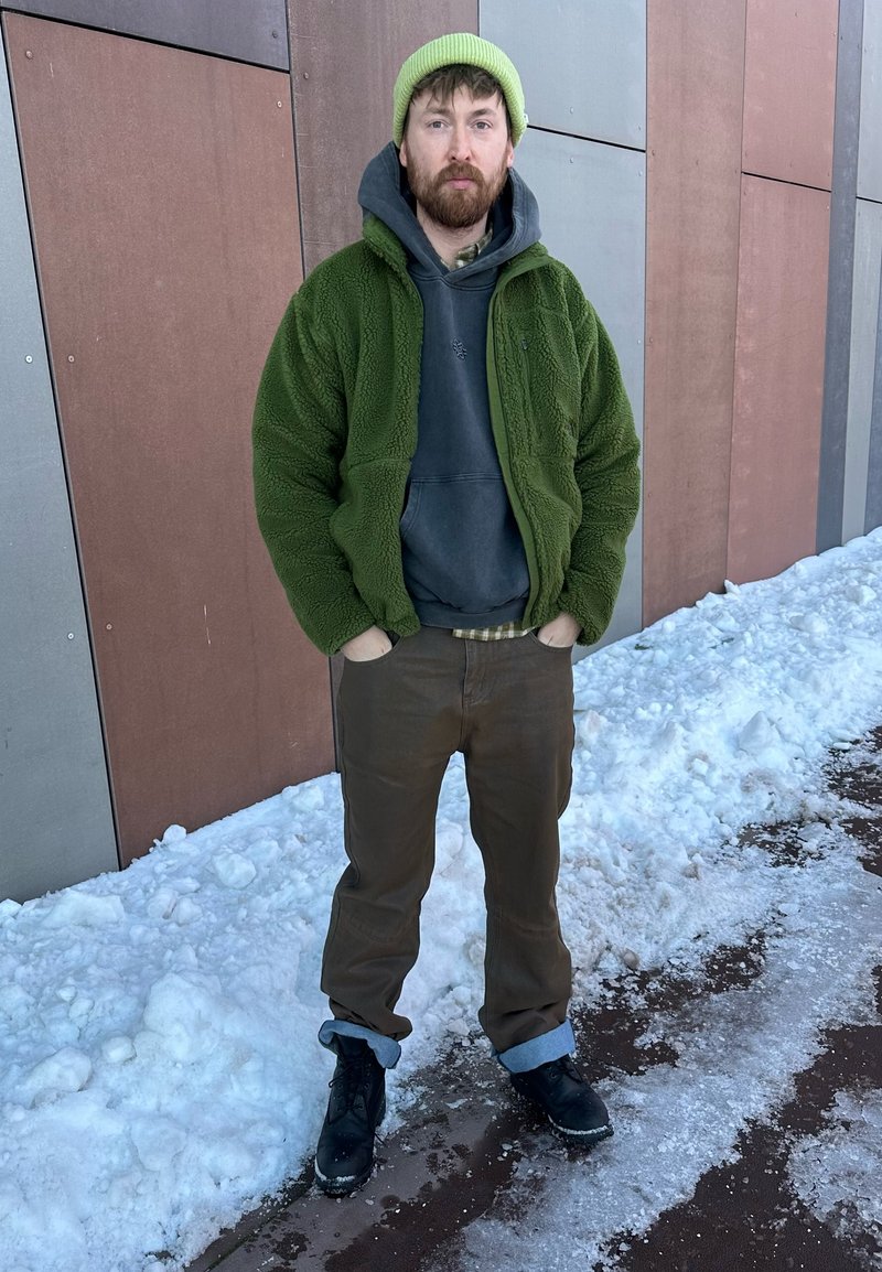 Mann mit grünem Mützchen, grüner Fleecejacke, dunklem Kapuzenpullover, brauner Hose und schwarzen Stiefeln steht mit den Händen in den Taschen auf verschneitem Boden neben einem Gebäude.