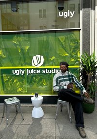 Una persona è seduta all'esterno dello "Ugly Juice Studio", accanto a uno sgabello bianco a clessidra e a una sedia di metallo. Lo sfondo presenta una plastica verde brillante e dei segnali.