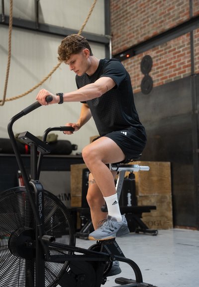 Bicicleta de aire negra con un ventilador grande y un asiento ajustable, siendo utilizada por una persona vestida con ropa atlética negra y zapatos grises, en el suelo de un gimnasio.