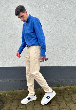Camisa de manga larga azul claro, pantalones crema y zapatillas blancas con detalles azules. Sosteniendo gafas de sol, de pie contra una pared texturizada.