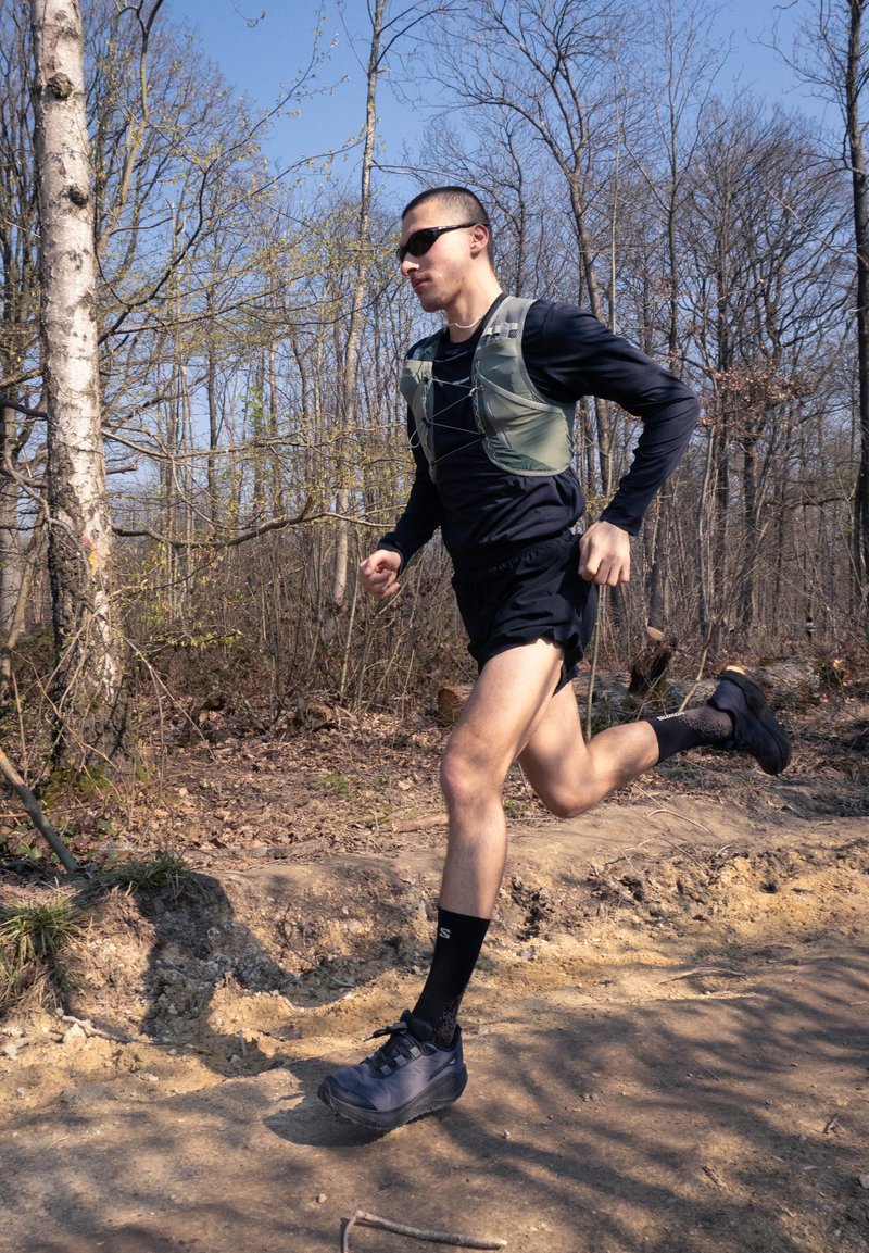 Man wearing sunglasses and athletic gear running on a dirt trail in a leafless forest during daytime.