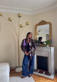 Veste en cuir marron, sac violet, écharpe à carreaux, jeans denim à jambes larges, chaussures marron ; décor moderne avec une lampe sur pied dorée haute et un miroir.
