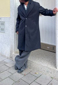 Czarny wełniany płaszcz o dopasowanym kroju, z dwoma guzikami i bocznymi kieszeniami, zestawiony z luźnymi ciemnymi dżinsowymi spodniami i czarnymi butami. Obok ściany.