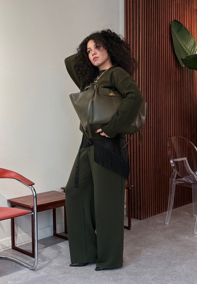 Mujer con cabello rizado, vestida con un conjunto verde oscuro, sosteniendo un bolso grande verde oscuro, de pie en una habitación moderna con pared de madera y silla transparente.