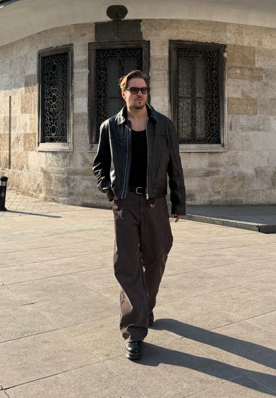 Chaqueta de cuero negra, camiseta sin mangas negra, pantalones anchos marrones y botas negras. Un hombre camina sobre una superficie pavimentada cerca de un edificio con ventanas decorativas.