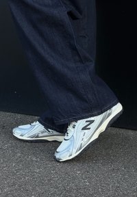 Pieds portant des baskets New Balance blanches et argentées avec des semelles noires, debout sur un asphalt gris à côté d'un mur noir, vêtus d'un pantalon bleu foncé.