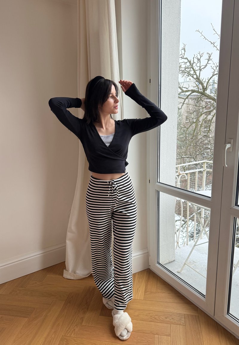 Femme en haut noir et pantalon rayé s'étirant près d'une fenêtre avec des rideaux blancs, regardant dehors une scène hivernale sur le balcon.