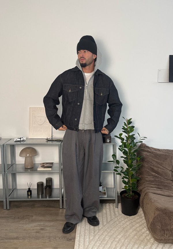 Homme portant un bonnet noir, une veste en denim noire par-dessus un sweat à capuche gris, un pantalon gris ample, debout dans une pièce moderne avec des étagères métalliques et une plante en pot.