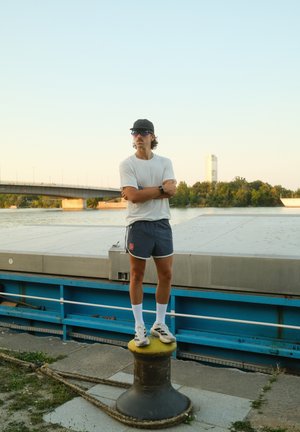 Pantalones cortos deportivos en azul marino con accents blancos, combinados con una camiseta blanca sencilla. De pie sobre un bolardo negro, usando gafas de sol y zapatillas.