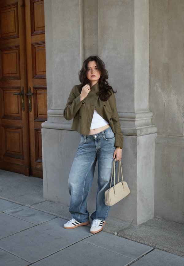Woman standing against a stone pillar wearing a cropped olive jacket, white top, loose jeans, white sneakers, and holding a beige handbag.