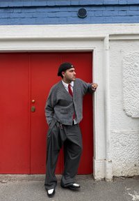 Uomo che indossa un cardigan grigio, cravatta rossa, pantaloni a righe sottili e basco nero, appoggiato a un muro bianco accanto a porte rosse su una strada.