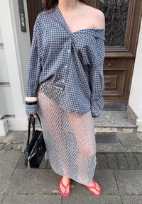 Persona con una camisa de cuadros de gran tamaño y un hombro descubierto, una falda de malla plateada sobre pantalones cortos negros, chanclas rojas, y sosteniendo un bolso negro.