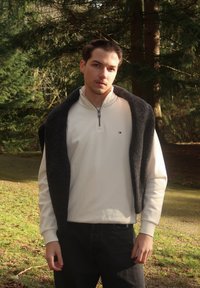 Νεαρός άνδρας που φοράει λευκό φούτερ με φερμουάρ μέχρι τη μέση και σκούρο πουλόβερ περασμένο στους ώμους, στέκεται σε εξωτερικό χώρο με δέντρα και γρασίδι στο υπόβαθρο.