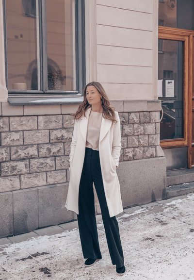 Abrigo largo blanco sobre una blusa clara y pantalones negros, con un cuello clásico y bolsillos laterales prominentes. Texturas suaves y ajuste entallado.