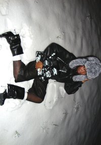 Personne allongée dans la neige portant une tenue en cuir noire, des bottes noires, un chapeau en fourrure grise et tenant un sac à main graphique noir.