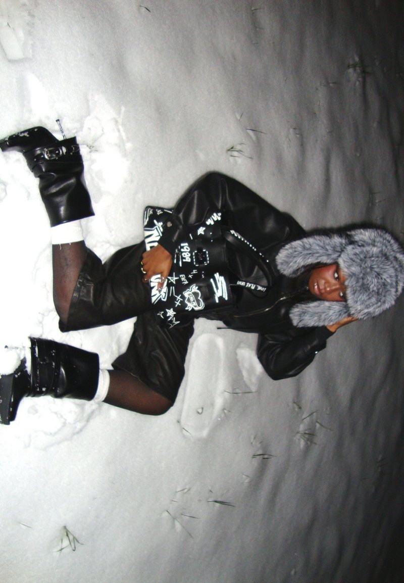 Personne allongée dans la neige portant une tenue en cuir noire, des bottes noires, un chapeau en fourrure grise et tenant un sac à main graphique noir.