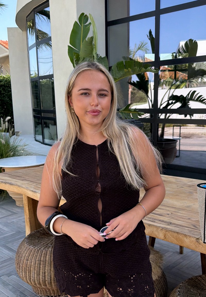Jeune femme aux longs cheveux blonds portant une tenue noire sans manches, debout près d'une table en bois à l'extérieur d'un bâtiment moderne avec des plantes.