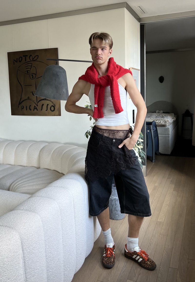Young man in a white tank top, red scarf, black shorts, and leopard print shoes poses with hand on hip in a modern living room.