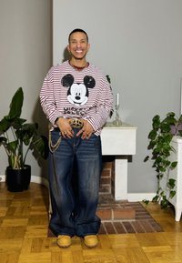 Ilgas dryžuotas marškinėlis su Mikio Pelės atvaizdu, tamsiai mėlyni plačios kelnės džinsai, šviesaus rudo atspalvio batai ir auksinės grandinėlės aksesuarai, esančio namų aplinkoje.