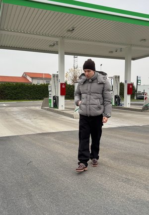 Chaqueta de plumas gris con capucha, pantalones negros y zapatos marrones, caminando por el pavimento de una gasolinera bajo un cielo nublado.
