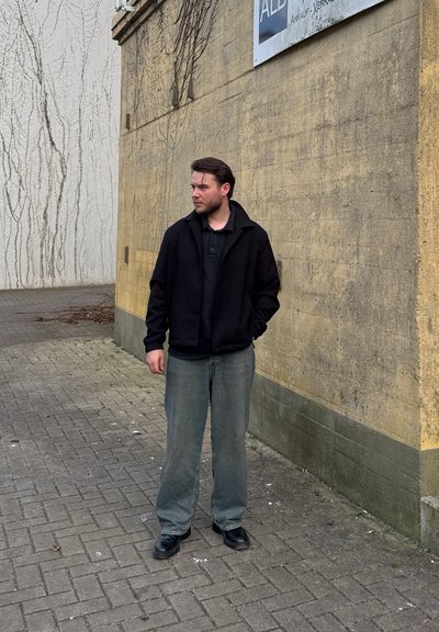 Chaqueta negra sobre un polo oscuro, pantalones de pierna ancha gris claro y zapatos negros. El fondo presenta una pared amarilla texturizada y un suelo pavimentado.
