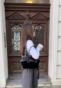 Geantă neagră din piele cu două mânere superioare, formă dreptunghiulară și accesorii argintii, poziționată în fața unei uși din lemn cu detalii elaborate.