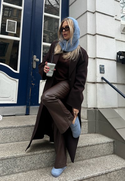 Pantalones de cuero marrón, abrigo marrón oversized, bufanda de punto azul clara y zapatos azules, sosteniendo un vaso en escaleras exteriores junto a una puerta azul.