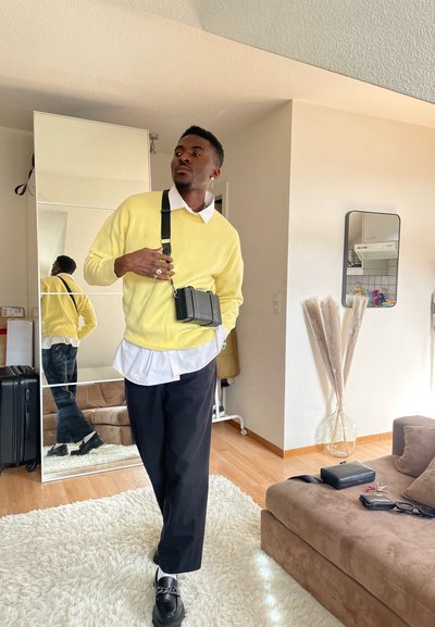 Suéter amarillo, camisa blanca debajo, pantalones negros y zapatos negros. Bolsa cruzada con herrajes. La reflección en el espejo muestra una vista parcial.