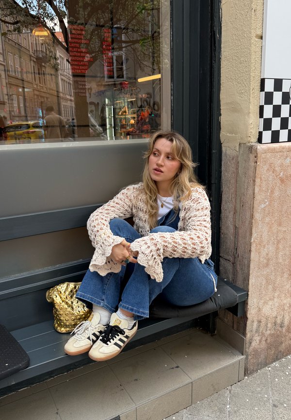 Ung kvinde med blond hår iført en hvid hæklet sweater og jeans, siddende på en bænk udenfor en café med en gylden nittebesat taske ved siden af sig.