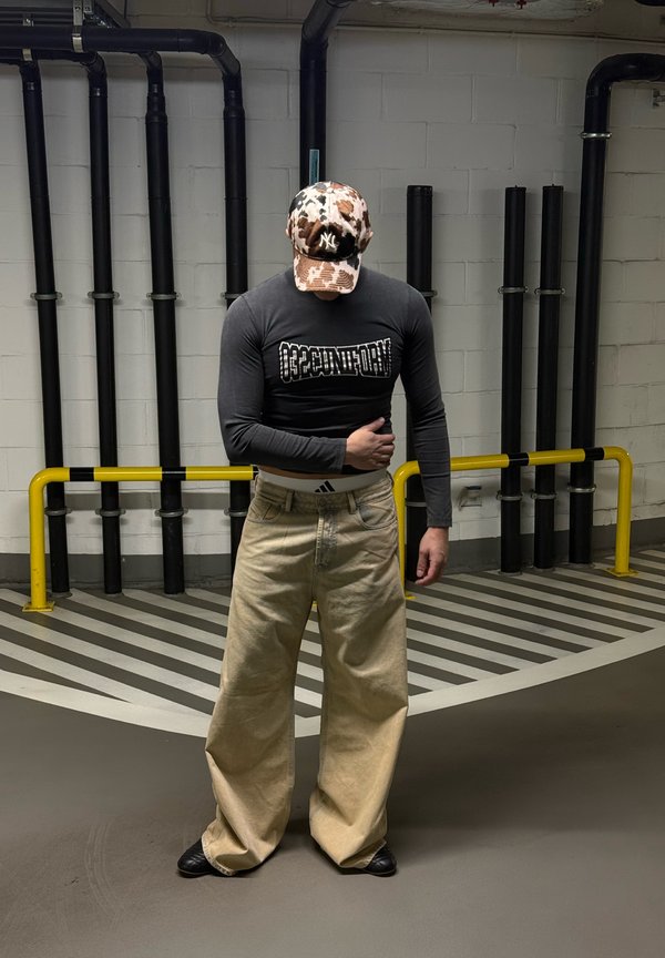 Bărbat cu șapcă de baseball camuflaj, cămașă neagră cu mâneci lungi și pantaloni largi bej stă într-un spațiu industrial cu podea în dungi și țevile negre.