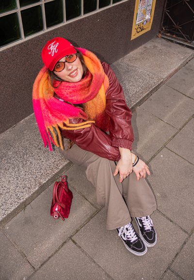 Gorra de béisbol roja, bufanda naranja y rosa, chaqueta de cuero marrón, pantalones beige, zapatillas negras. Pequeño bolso rojo en el suelo al lado.