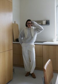 Pull gris duveteux avec col blanc, pantalon blanc, lunettes rondes, debout dans une cuisine avec des meubles en bois et une décoration minimaliste.