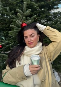 Cappotto beige con bottoni dorati, sciarpa crema, che tiene una tazza a strisce verdi. Capelli lunghi e scuri, accenti rossi con albero di Natale sullo sfondo.