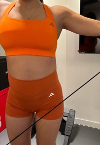 Personne portant un brassard de sport orange et un short avec des logos Adidas, s'entraînant avec un câble de résistance dans une salle de sport à domicile.
