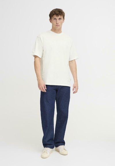 Camiseta de algodón beige de manga corta, combinada con pantalones de mezclilla azul oscuro y zapatillas de color claro. Diseño sencillo y recto.