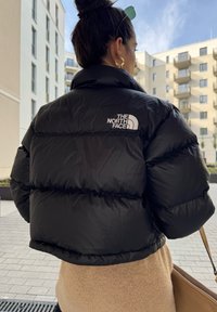 Μαύρο μπουφάν puffer με ψηλό γιακά, διαθέτει ραμμένα οριζόντια τμήματα και λευκή επωνυμία στο επάνω μέρος της πλάτης. Λείο, γυαλιστερό υλικό.