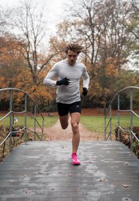 Futó világos szürke hosszú ujjú felsőben és fekete rövidnadrágban egy fapallós hídon, fekete kesztyűt és élénk rózsaszín futócipőt visel. Őszi háttér.