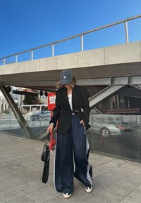 Donna che indossa un blazer nero, una maglietta bianca, pantaloni sportivi blu navy larghi con strisce bianche, sneakers e un cappellino blu, mentre tiene una borsa nera all'aperto.