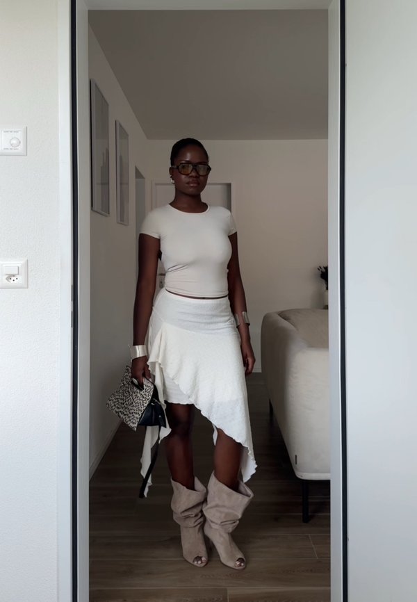Woman wearing glasses, a fitted white top, asymmetrical white skirt, slouchy beige boots, and holding a patterned handbag indoors.