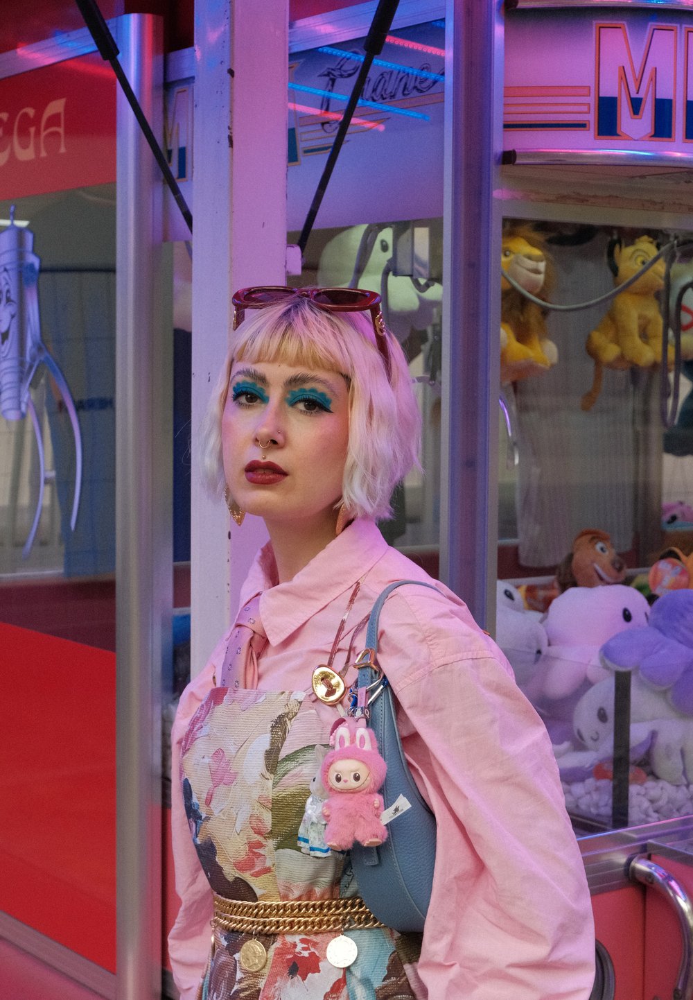 Blonder Bob-Haarschnitt; pinkes Hemd; blumengemusterte Schürze; goldene Kettengürtel; blaue Handtasche mit plüschigen Anhängern; lebendiger Hintergrund mit Plüschspielzeugen.