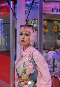 Blonder Bob-Haarschnitt; pinkes Hemd; blumengemusterte Schürze; goldene Kettengürtel; blaue Handtasche mit plüschigen Anhängern; lebendiger Hintergrund mit Plüschspielzeugen.